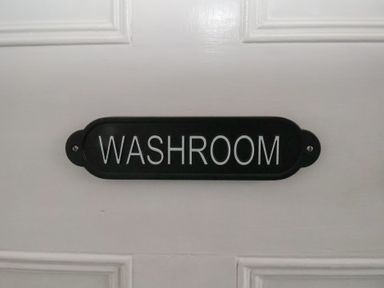 A black sign with white letters saying washroom mounted on a white wooden door at Trackside in Ruswarp