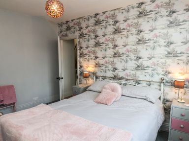 A bedroom with a double bed pillows and blankets two bedside tables with lamps wallpaper on one wall door open to another room at Trackside in Ruswarp