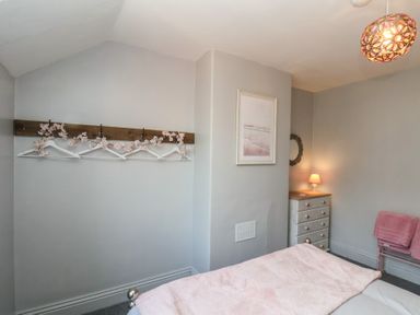 A bedroom with a bed covered in a pink blanket a wooden dresser with a lamp a mirror a framed picture a wall-mounted coat rack with hangers and pink flowers and a pink towel on a rack at Trackside in Ruswarp