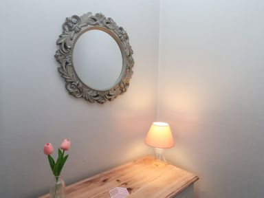 A corner of a room with a wooden table holding a vase with two pink tulips and a lit lamp below an ornate round mirror at Trackside in Ruswarp