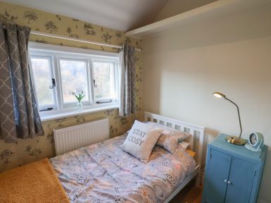 A bedroom with a single bed, patterned bedding, a pillow with stay cosy text, a blue bedside cabinet with a lamp and clock and a window with curtains at Trackside in Ruswarp
