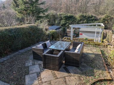 An outdoor patio area with wicker furniture and cushions surrounded by hedges and trees at Trackside in Ruswarp