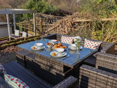 An outdoor patio with a glass table set with bowls plates croissants and fruit surrounded by wicker chairs with cushions at Trackside Ruswarp