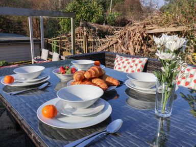 An outdoor dining table set with white bowls plates spoons croissants strawberries mandarins and a glass vase with white flowers at Trackside Ruswarp