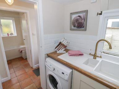 A kitchen area with a washing machine a sink with a brass faucet a knife set on the counter and a doorway leading to a bathroom with a bathtub and a window at Cherry Blossom Cottage in Stratford-Upon-Avon