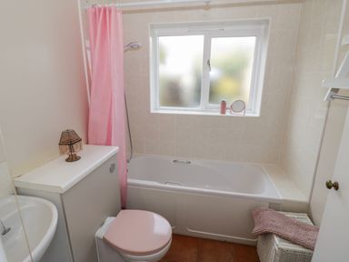 A bathroom with a bathtub shower with a pink curtain a toilet with a pink lid a small sink a window a wicker basket and a small lamp at Cherry Blossom Cottage in Stratford-Upon-Avon