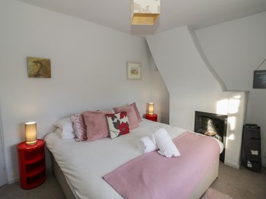 A bedroom with a double bed with pink pillows and a pink bed runner between two red bedside tables with lamps and a fireplace at Cherry Blossom Cottage in Stratford-Upon-Avon