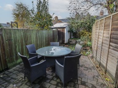 An outdoor patio with a round glass table and five wicker chairs surrounded by wooden fences at Cherry Blossom Cottage in Stratford-Upon-Avon