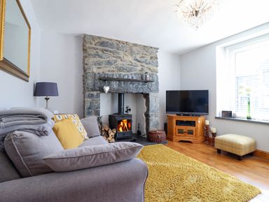 A living room with a stone fireplace wood stove grey sofa yellow rug and a television at Dola in Pentre Du near Betws-Y-Coed