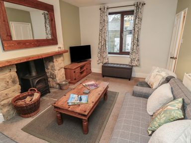 A living room with a wooden coffee table magazines on it a gray sofa cushions a fireplace with a wood-burning stove a wicker basket with logs a wooden TV cabinet a window with floral curtains and a gray ottoman at Providence Cottage in Ugglebarnby near Sleights