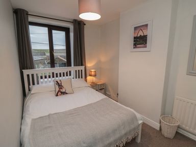 A bedroom with a bed a bedside table with a lamp a window with curtains a framed picture and a laundry basket at Providence Cottage in Ugglebarnby near Sleights