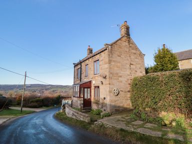 A stone cottage on a curved road with a garden and hills in the background at Providence Cottage in Ugglebarnby near Sleights