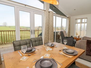 A dining table set with plates bowls and glasses near windows overlooking a wooden deck and green fields at Seren Lodge in Builth Wells
