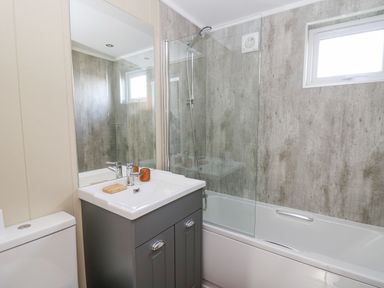 A bathroom with a toilet, a sink with cabinet, a mirror, a bathtub with a glass shower screen and a window at Seren Lodge in Builth Wells