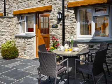 An outdoor patio with a round table set with cups and juice and four wicker chairs in front of a stone wall house with wooden door and two windows at Teifi Cottage in Cardigan Near Cenarth