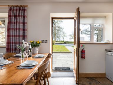 A dining area with a wooden table set with plates glasses flowers and an open door showing an outdoor patio at Teifi Cottage Cardigan Near Cenarth
