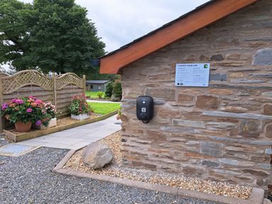 An outdoor wall with an electric car charging station and a sign next to a garden with flowers and a wooden fence at Teifi Cottage in Cardigan Near Cenarth