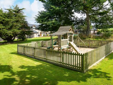 An outdoor playground with a wooden play structure surrounded by a wooden fence and grass at Teifi Cottage in Cardigan Near Cenarth