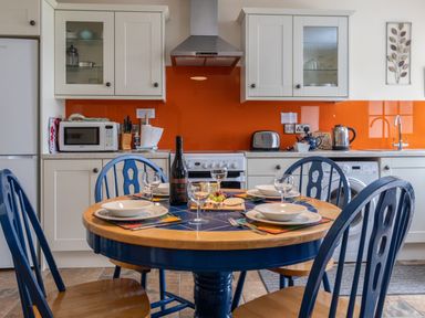 A kitchen with a round dining table set for four with blue chairs and kitchen cabinets with orange backsplash at Cych Cottage Cardigan Near Cenarth