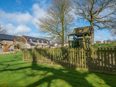 A fenced playground with a wooden play structure and slides near stone cottages at Cych Cottage Cardigan Near Cenarth