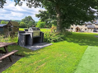 A garden with a built barbecue grill a wooden picnic table a large tree and a stone house in the background at Towy Cottage in Cardigan Near Cenarth