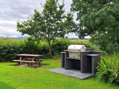 An outdoor area with a wooden picnic table and a built-in brick barbecue grill surrounded by trees and grass at Towy Cottage Cardigan Near Cenarth