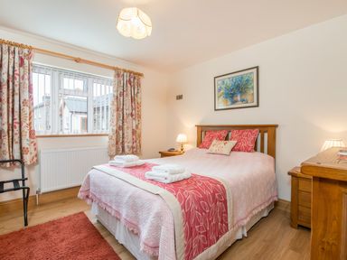 A bedroom with a double bed with pink bedding pillows and folded towels a window with floral curtains bedside tables with lamps a painting on the wall and wooden furniture at Gwaun Cottage in Cardigan Near Cenarth