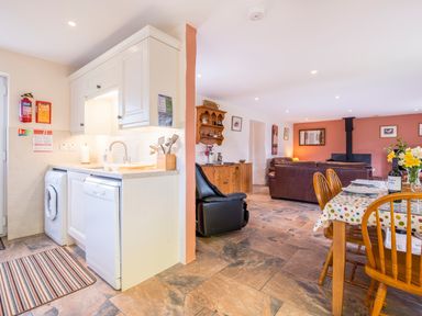 A kitchen with white cabinets and appliances next to a dining area with a wooden table and chairs and a living room with leather sofas and a wood stove at Cothi Cottage in Cardigan Near Cenarth