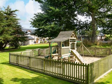 A fenced playground with a wooden play structure on grass at Cothi Cottage in Cardigan Near Cenarth