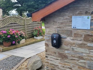 A stone wall with an electric car charger and instructions next to a garden with flowers and a wooden fence at Hirwaun Cottage Cardigan Near Cenarth
