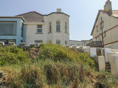 A two story white house with bay windows behind a white fence and overgrown grass at The Harbour in Rhosneigr