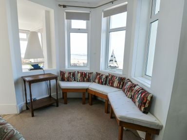 A corner seating area with white cushions and patterned pillows next to windows with a wooden side table and a model sailboat on the windowsill at The Harbour in Rhosneigr