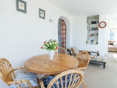 A dining area with a round wooden table surrounded by four wicker chairs with cushions and flowers in a vase on the table at The Harbour in Rhosneigr