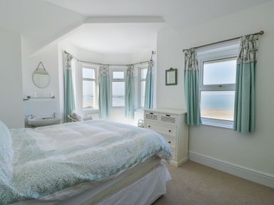 A bedroom with a bed a chest of drawers a bay window with curtains and an ocean view at The Harbour in Rhosneigr