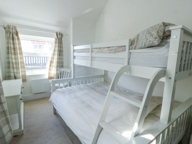 A bedroom with a white bunk bed a white chair a window with plaid curtains and a small white cabinet at The Harbour in Rhosneigr