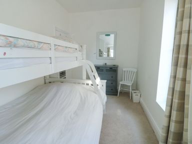 A bedroom with a white bunk bed a dresser a chair and a window with plaid curtains at The Harbour in Rhosneigr