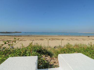 A sandy beach with calm sea and clear sky viewed over a white wall with greenery at The Harbour in Rhosneigr