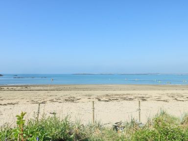 A sandy beach with some plants and a rope fence in the foreground and a calm sea with kayakers in the background at The Harbour in Rhosneigr