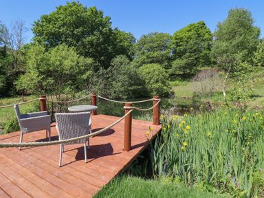 A wooden deck with two chairs and a table overlooking a pond with trees and yellow flowers at Celyn in Bontddu near Dolgellau