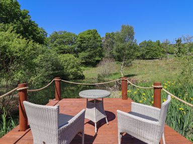 An outdoor wooden deck with two wicker chairs and a round table overlooking a pond surrounded by trees and greenery at Celyn in Bontddu near Dolgellau