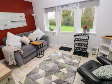 A sitting room with a gray sofa with cushions a small wooden side table a patterned rug a black leather chair a shoe rack and a door leading to a garden at Orchard View in Pembroke