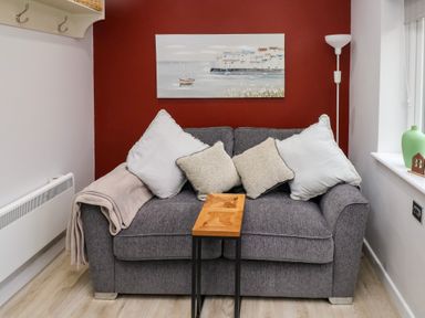 A gray sofa with four cushions and a beige throw blanket a small wooden table a floor lamp a painting on a red wall a window with a green vase and a small wooden house ornament at Orchard View in Pembroke