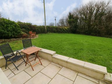 A patio with two black chairs and a wooden table overlooking a lawn with a bench and trees in the background at Orchard View in Pembroke