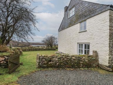 The exterior of a stone house with white walls and slate roof next to a gravel area and a small stone wall with a wooden gate opening to a grassy yard with trees and a distant view of water at Poldue House Watergate near Camelford