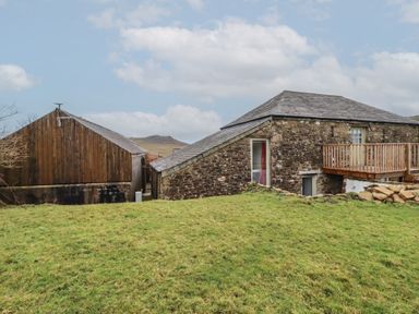 A stone house with a wooden balcony and a wooden barn in a grassy area at Logan Barn in Poldue near Camelford
