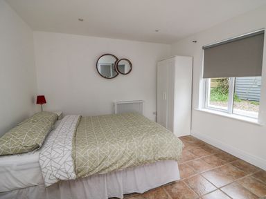 A bedroom with a bed with patterned bedding a white wardrobe two round mirrors a red lamp and a window with a blind at Bayside Cottage in Rosslare Strand County Wexford
