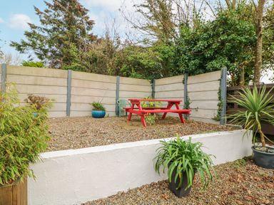 An outdoor area with a red picnic table, green plastic chair, potted plants, and a concrete panel fence at Bayside Cottage in Rosslare Strand County Wexford