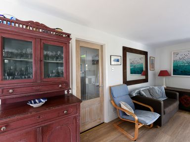 A room with a red cabinet filled with glassware a wooden framed glass door a blue cushioned chair brown sofa with cushions a mirror and framed pictures on the wall and a table lamp at Northern Lights in Polzeath