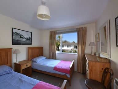 A bedroom with two single beds a nightstand a lamp a dresser a chair and a window with curtains at Northern Lights in Polzeath