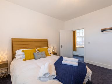 A bedroom with a bed with mustard headboard yellow and grey cushions folded towels on navy bedspread and open door showing window with mustard curtains at Groves House Castletown near Fortuneswell